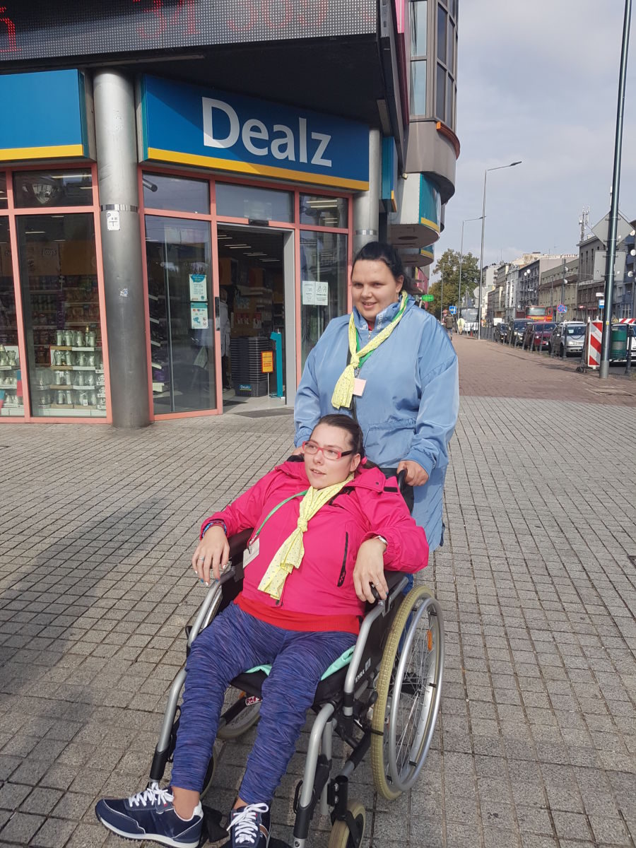 Dwie mieszkanki pozują do zdjęcia na tle ulicy, jedna z nich siedzi na wózku inwalidzkim, druga trzyma rączki wózka