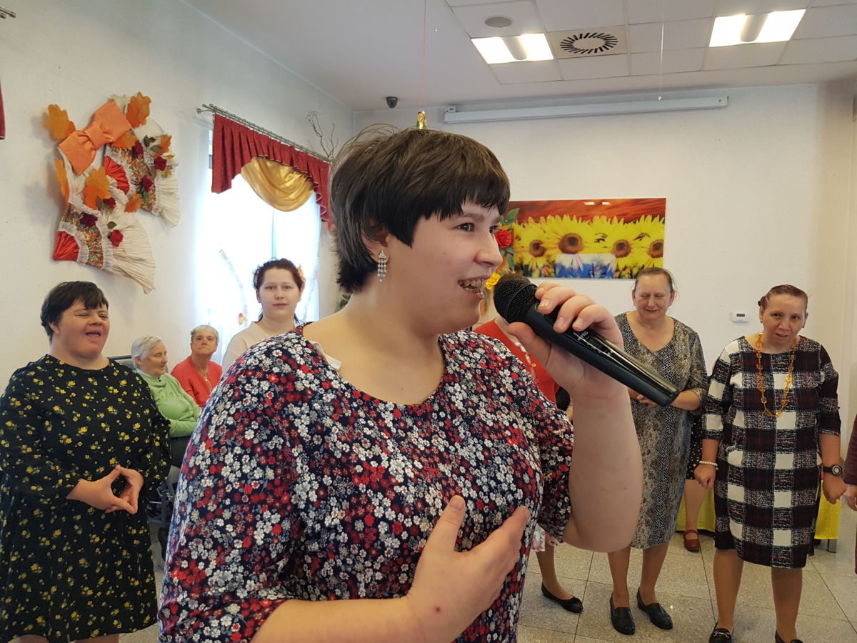 Uśmiechnięta mieszkanka śpiewa do mikrofonu, w tle kilka mieszkanek bioracych udział w zabawie na sali integracyjno - terapeutycznej