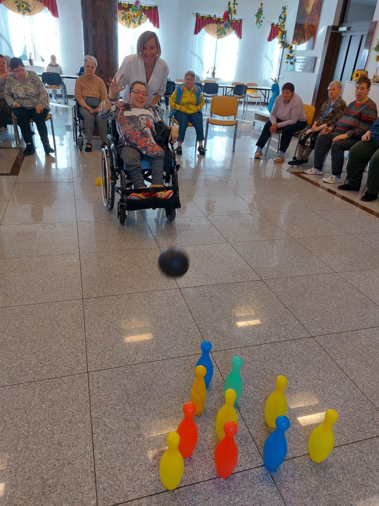 Roześmiana mieszkanka na wózku inwalidzkim grająca w kręgle, dopingowana przez opiekunkę. W tle siedzące współzawodniczki. 