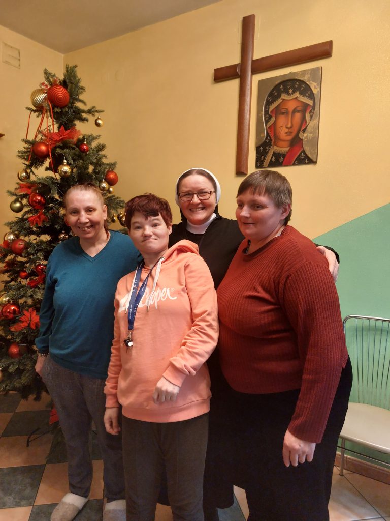 Trzy uśmiechnięte mieszkanki pozują do zdjęcia z Siostrą Nazaretanką - w tle choinka oraz wiszący krzyż i obraz Maryi