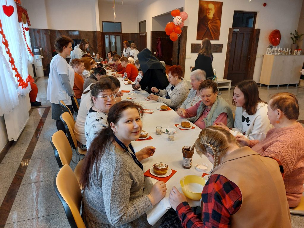 Mieszkanki wraz z wolontariuszami siedzą przy długim stole na sali integracyjno - terapeutyczneji dekorują pączki, w tle kilka osób stoi przy stole