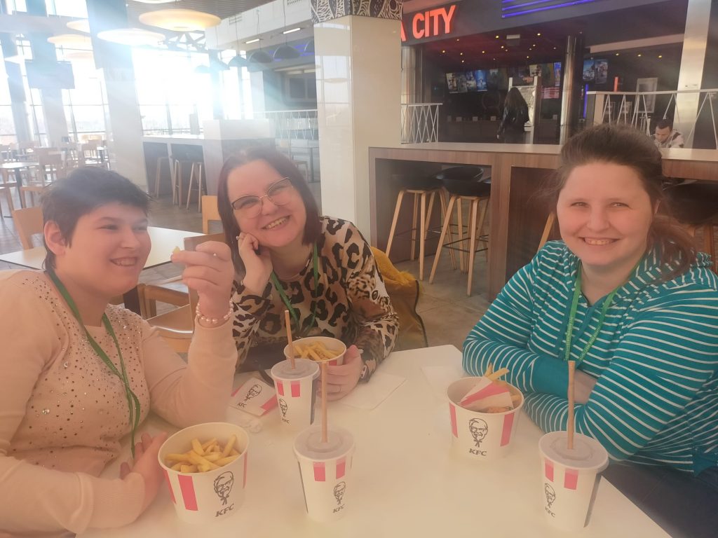 Trzy uśmiechnięte mieszkanki siedzą przy stoliku, na którym są zestawy fast food
