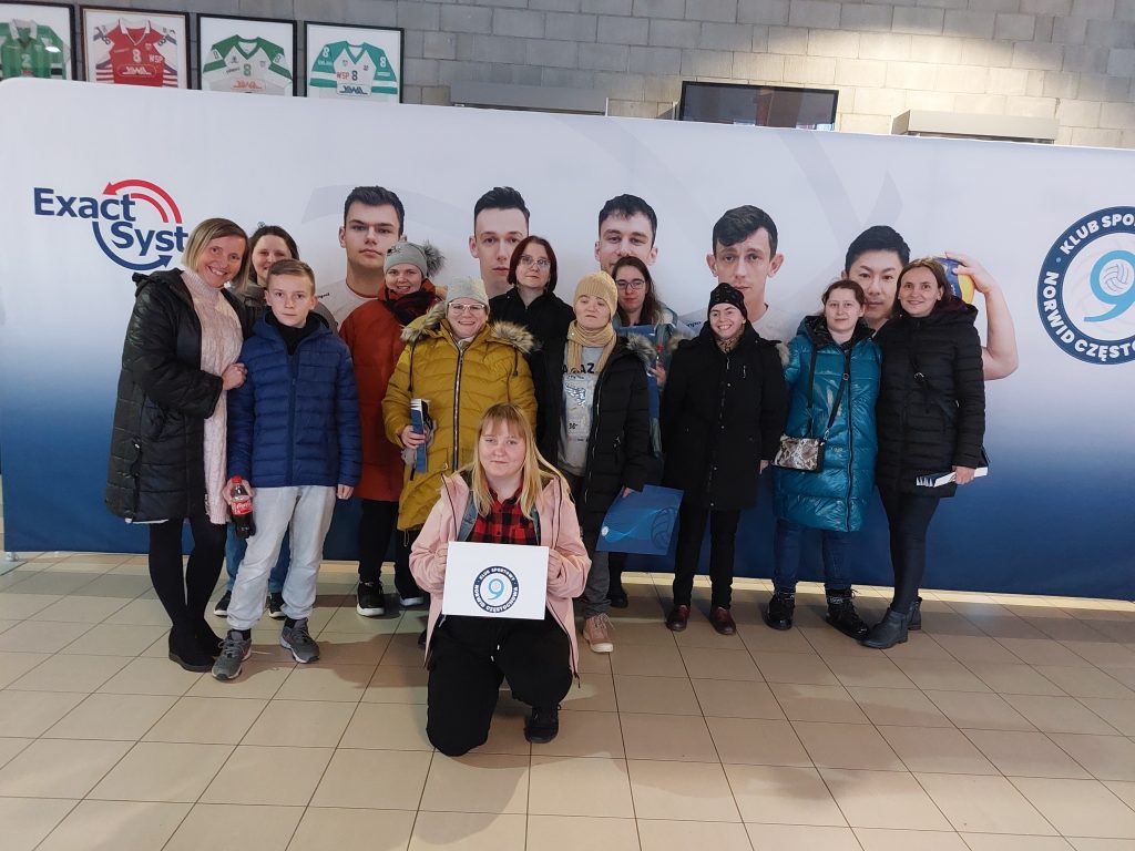 Zdjęcie grupowe uśmiechniętych mieszkanek z opiekunkami na tle plakatu z drużyną z Częstochowy,  przed grupą kuca jedna mieszkanka z plakatem drużyny w ręku