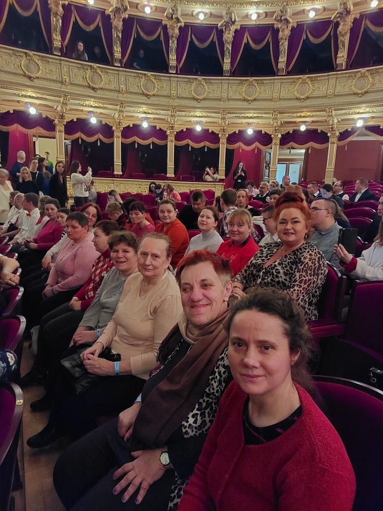 Uśmiechnięte osoby z zespółu Michalinki wraz z jedną z pań odpowidzialnych za grupę siedzące na widowni teatru