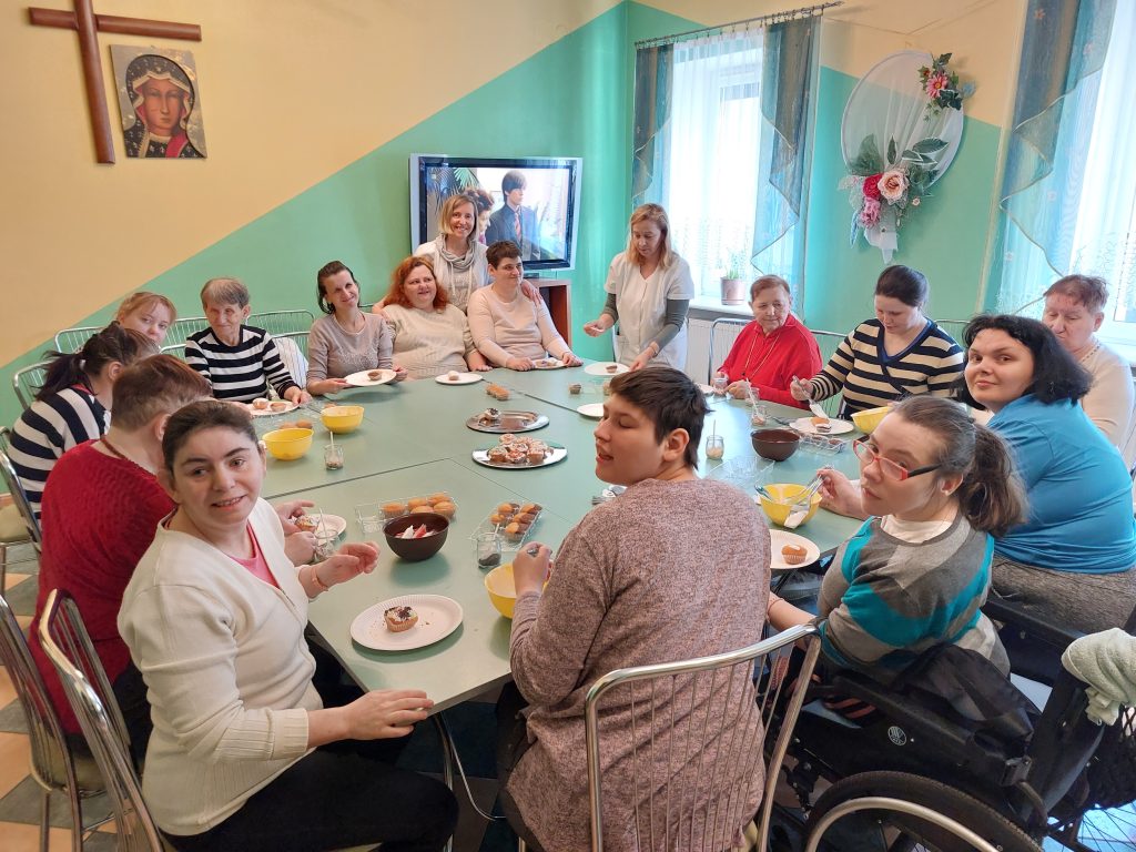 Wspólne zdjęcie mieszkanek siedzących przy stole przygotowujących babeczki w kawiarni domu, w tle stojące terapeutki