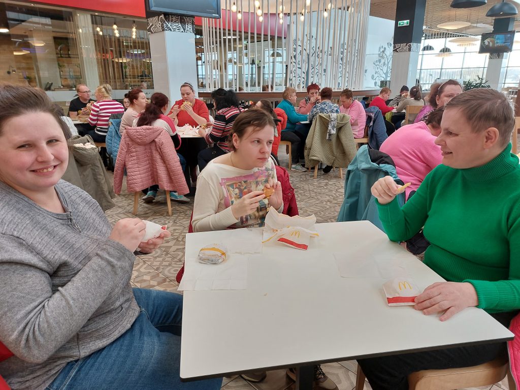 Trzy mieszkanki siedzą przy stole i pozują do zdjęcia podczas jedzenia w Mc Donald's, w tle jedzące przy stolikach pozostałe mieszkanki