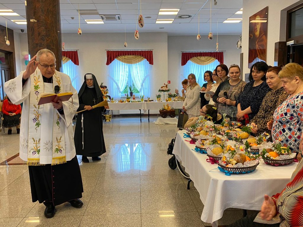 Ksiądz biskup odmawia modlitwę podczas święcenia pokarmów. Z prawej strony zebrane mieszkanki przy stole zastawionym koszyczkami do święcenia
