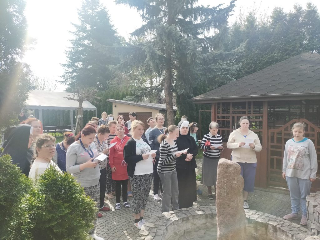 Grupa Mieszkanek wraz z Siostrami Albertynkami stoi zebrana na podwórzu Domu - śpiewają hymn,, część osób ma w rękach kartki z tekstem