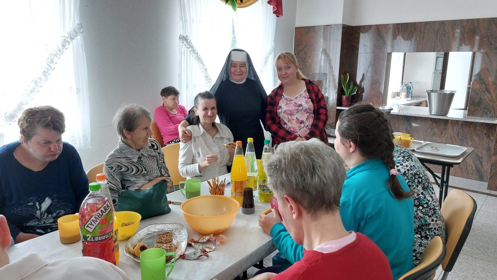 Uśmiechnięte Siostra Albertynka i mieszkanka pozują na stojąco do zdjęcia, pozostałe osoby siedzą przy stole podczas poczęstunku na sali integracyjno - terapeutycznej