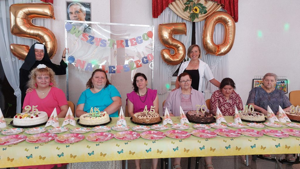 Sześć uśmiechniętych jubilatek siedzi przy stole zastawionym tortami i pozuje do zdjęcia ze stojącymi za nimi uśmiechniętą siostrą Albertynką i pracownikiem terapii