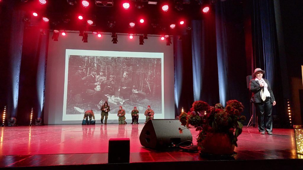 Zespół Michalinki na scenie podczas występu - po prawej stronie stoi wokalistka z mikrofonem w ręku, ma kapelusz na głowie, w tle są mieszkanki przebrane za żołnierzy, na ekranie za nimi prezentacja multimedialna obrazująca treść śpiewanej piosenki