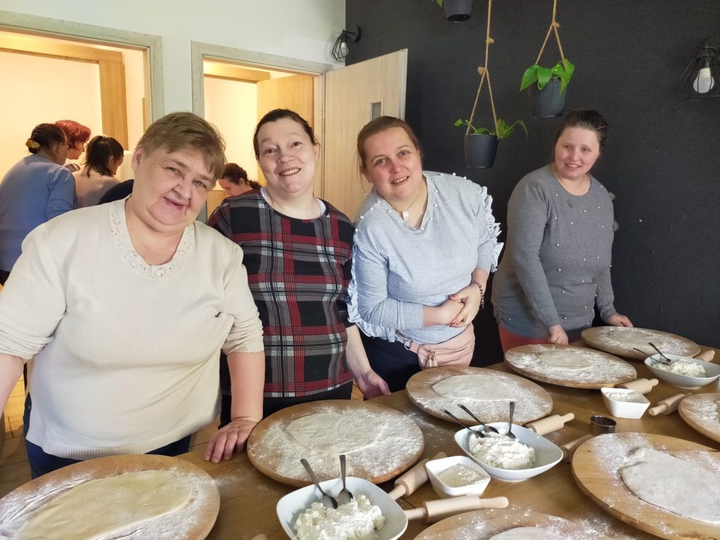 Cztery uśmiechnięte mieszkanki stojąc przy stole pozują do zdjęcia. Każda z nich ma przed sobą deskę, na której przygotowane jest ciasto na pierogi, obok jest farsz i wałki