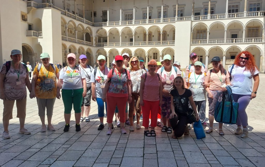 Uśmiechnięta grupa mieszkanek razem z opiekunkami pozuje do zdjęcia stojąc w krużgankach zamku na Wawelu, jedna osoba kuca przed grupą 