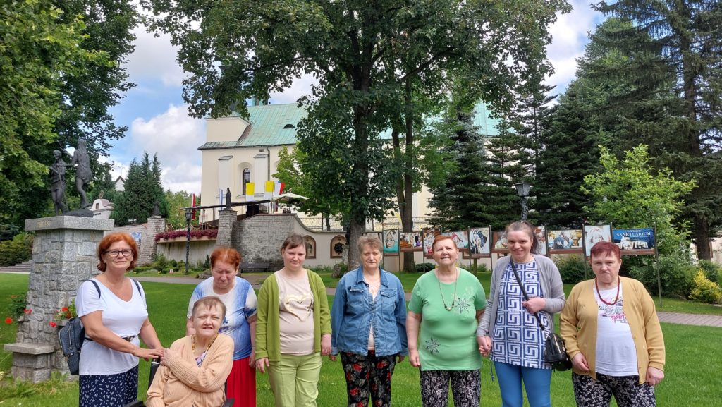 Uśmiechnięte mieszkanki wraz z opiekunką grupy stojąc pozują do zdjęcia na tle sanktuarium. Jedna osoba siedzi na wózku inwalidzkim