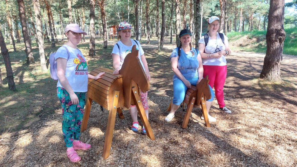 Roześmiane mieszkanki pozują do zdjęcia przy drewnianych konikach na terenie parku. Dwie osoby opierają ręce na większym koniu, trzy osoby stoją , czwarta siedzi na mniejszym koniu