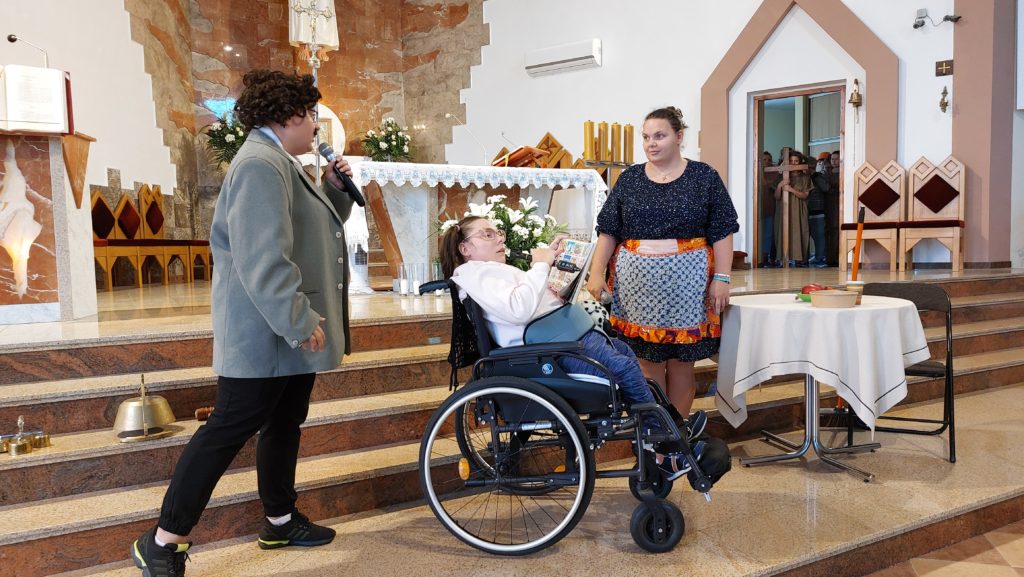 Trzy mieszkanki podczas występu w kościele. Dwie osoby stoją, jedna jest na wózku inwalidzkim, wszystkie mają w ręku mikrofony