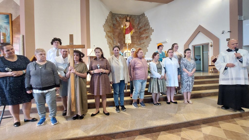 Osoby z Zespołu Michalinki stoją przed ołtarzem w kościele. Na pierwszym planie mieszkanka z krzyżem w ręku i w koronie cierniowej na głowie, obok stoi ksiądz proboszcz z mikrofonem 