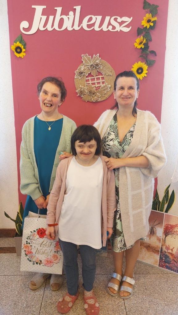 Dwie uśmiechnięte mieszkanki stojąc pozują do zdjęcia z jubilatką na tle ścianki foto z napisem jubileusz. Jubilatka trzyma w ręku torbę prezentową
