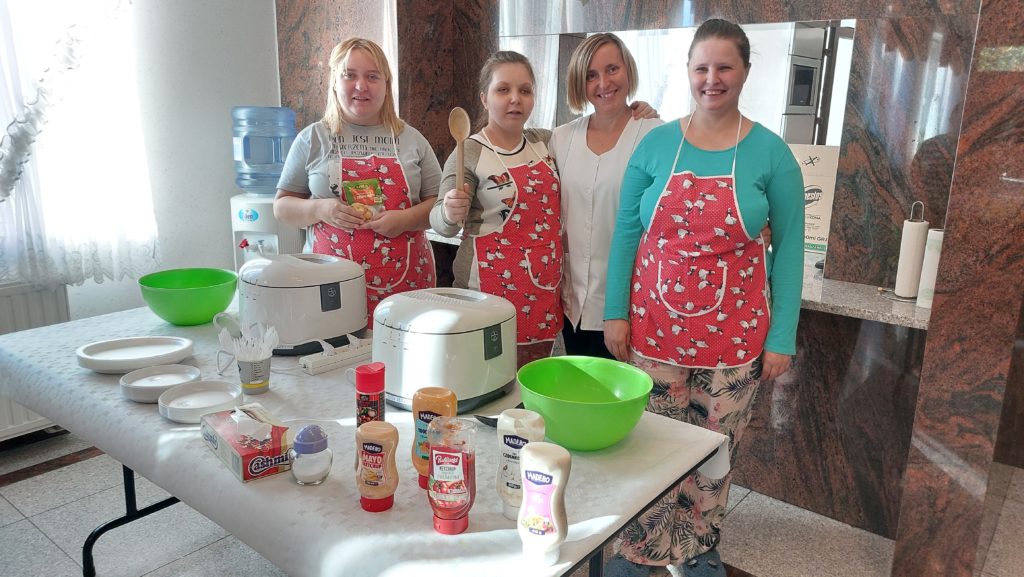 Trzy roześmiane mieszkanki stojąc pozują do zdjecia na sali integracyjno - terapeutycznej z jedną z osób z personelu. Na sobie mają fartuszki kuchenne przed nimi stoi stół, na stole akcesoria do robienia frytek