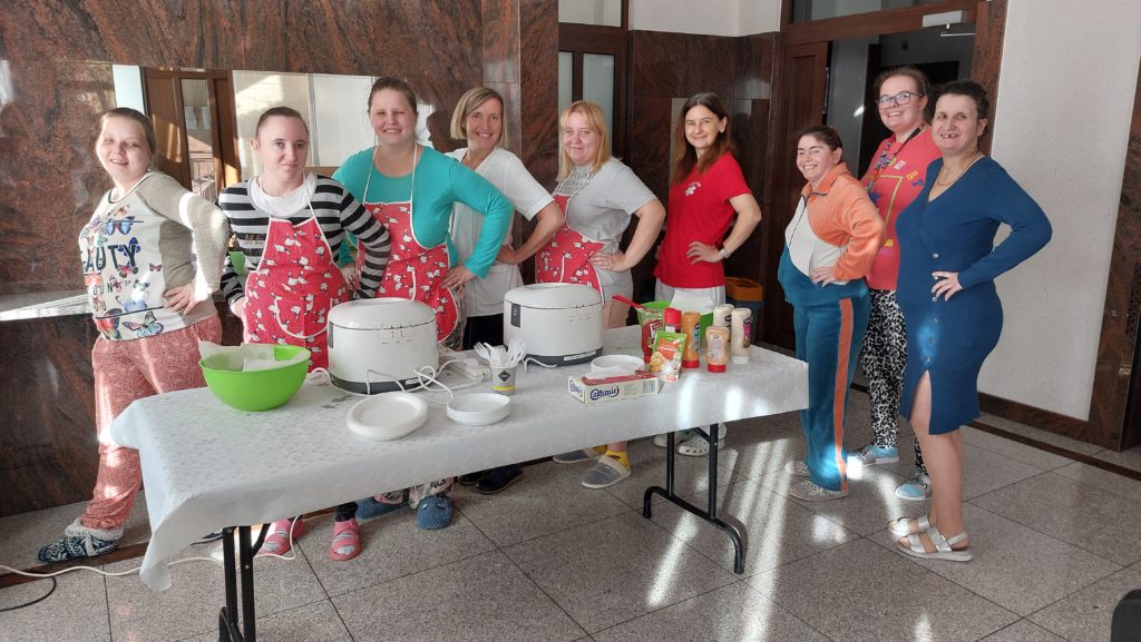 Grupa uśmiechniętych mieszkanek stojąc pozuje do zdjęcia na sali integracyjno - terapeutycznej z dwiema osobami z personelu. Więszośc osób stoi bokiem z ręką na biodrze, przed nimi stoi stół, leżą na nim akcesoria do robienia frytek