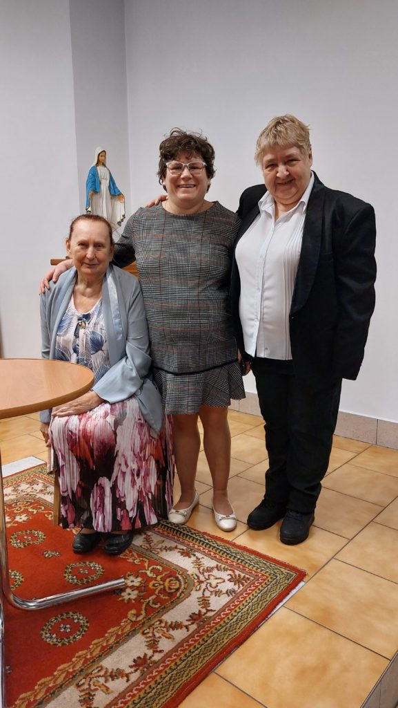 Trzy uśmiechnięte mieszkanki pozują do zdjęcia. Dwie z nich stoją, jedna z nich siedzi przy okrągłym stoliku, w tle figura Matki Bożej