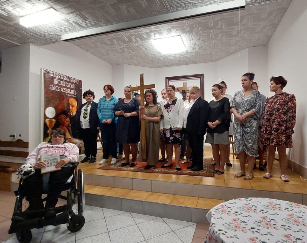 Zespół Michalinki stojąc pozuje do zdjęcia, pośrodku grupy stoi mieszaknka z krzyżem w rękach. Na pierwszym planie jest mieszkanka siedząca na wózku inwalidzkim