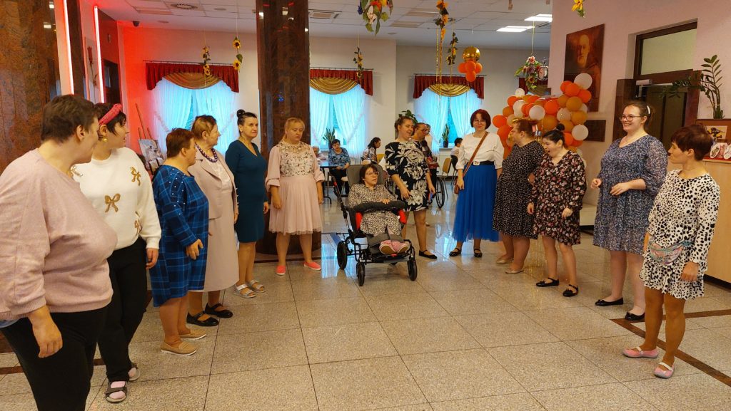 Grupa mieszkanek stoi ustawiona w półkole na sali integracyjno - terapeutycznej, wśród nich jest osoba siedząca na wózku inwalidzkim