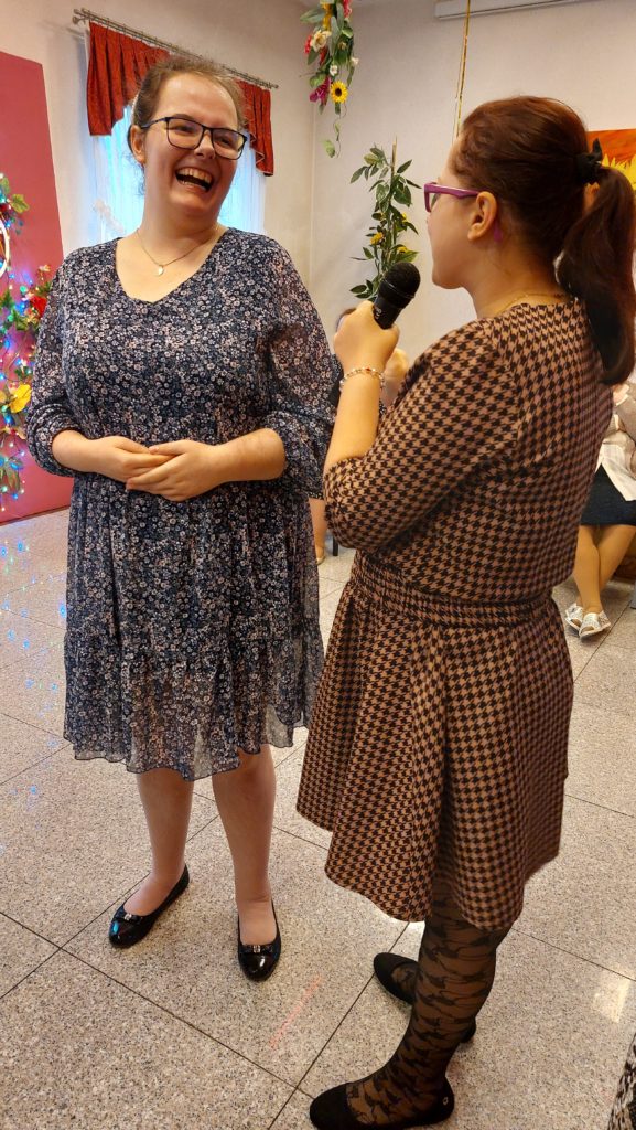 Roześmiana mieszkanka stoi wpatrując się w drugą mieszkankę, która ma w ręku mikrofon