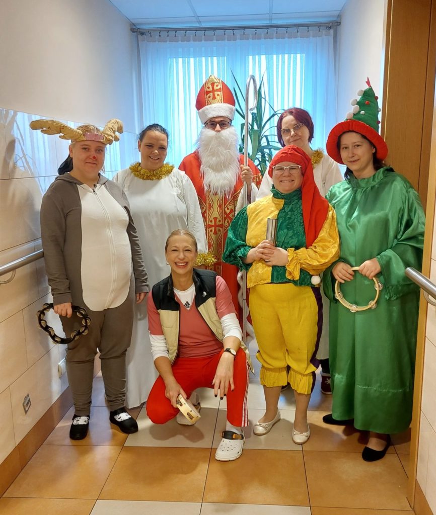 Uśmiechnięte mieszkanki przebrane za śnieżynki, łosia, wróżkę, elfa stojąc pozują do zdjęcia z Mikołajem i rehabilitantką na korytarzu Domu na trzecim piętrze, w ręku mają instrumenty. Roześmiana rehabilitantka kuca przed grupą z tamburynem w ręku