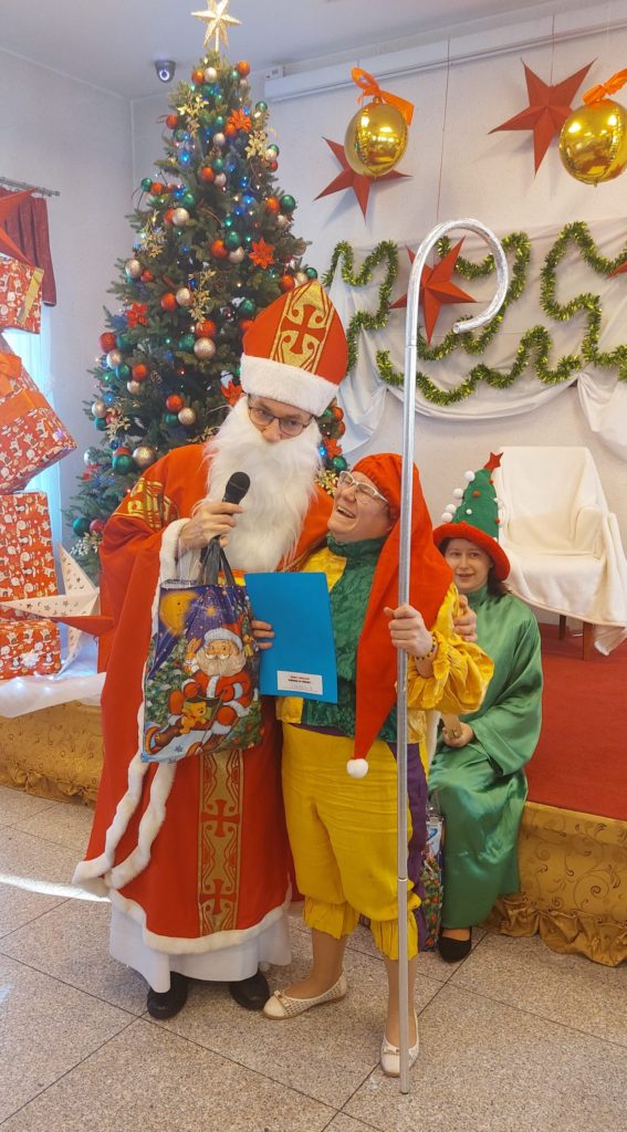 Mikołaj pozuje do zdjęcia z roześmianą mieszkanką przebraną za elfa na sali integracyjno - terapeutycznej, w tle na scenie siedzi mieszkanka w stroju wróżki. Mikołaj ma w ręku mikrofon i paczkę