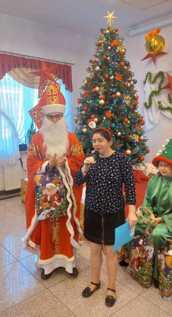 Mikołaj stojąc pozuje do zdjęcia z mieszkanką na sali integracyjno - terapeutycznej, w ręku ma paczkę, klaszcze. Mieszkanka ma w ręku mikrofon i kartkę, w tle na scenie stoi choinka, obok siedzi mieszkanka w stroju wróżki. 