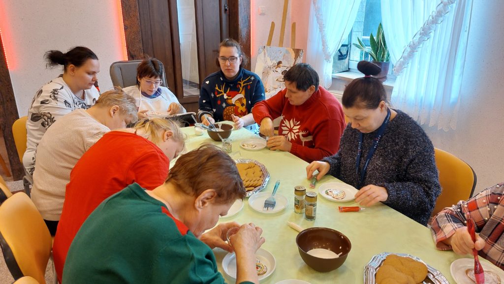 Grupa mieszkanek na sali integracyjno - terapeutycznej siedzi skupiona przy stołach dekorując pierniki