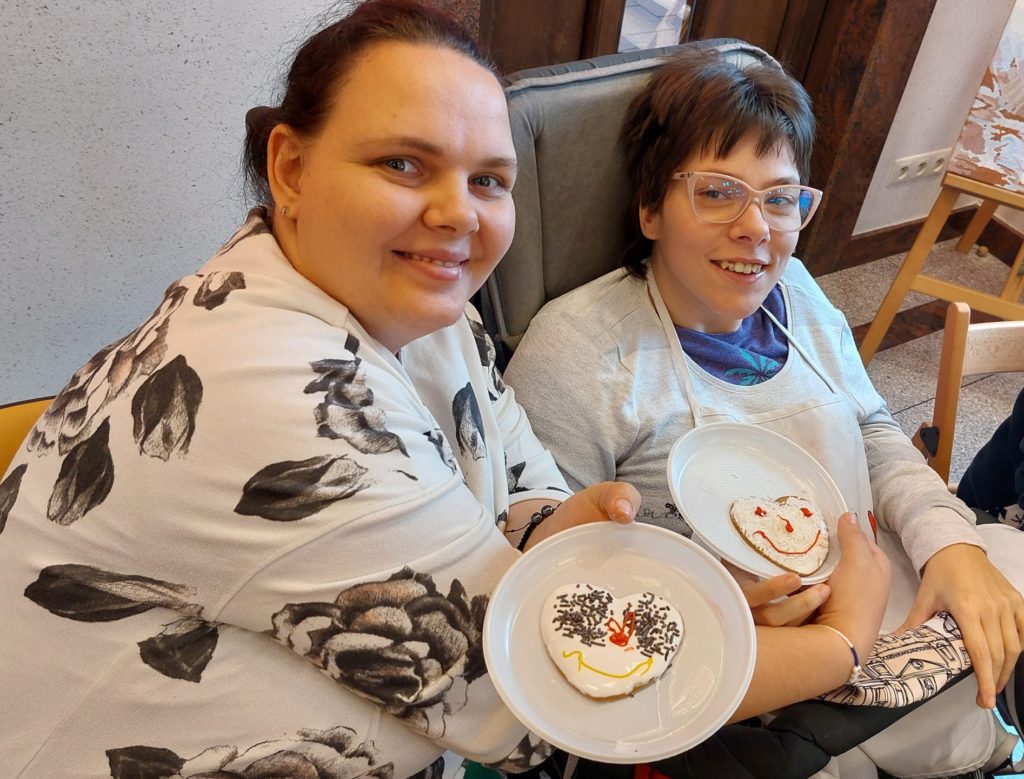 Dwie uśmiechnięte mieszkanki siedząc pozują do zdjęcia na sali integracyjno - terapeutycznej, prezentują telarze z udekorowanymi pierniczkami. Jedna z nich siedzi na wózku inwalidzkim