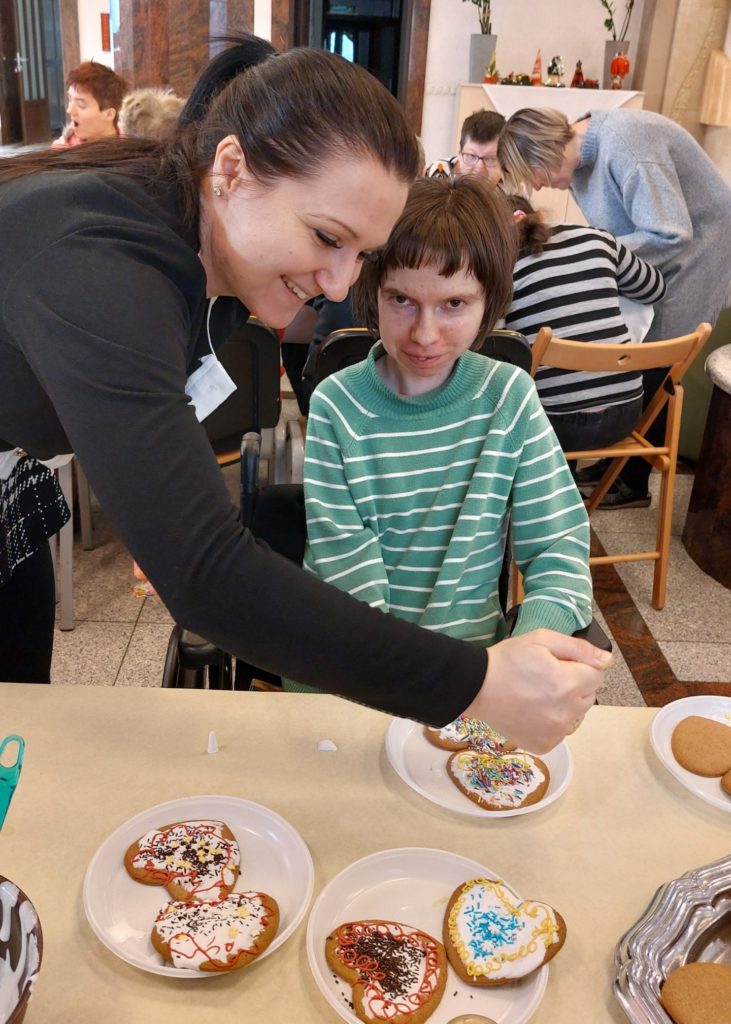 Roześmiana terapeutka na sali integracyjno - terapeutycznej pochyla się nad uśmiechniętą mieszkanką siedzącą przy stole na wózku inwalidzkim. Terapeutka dekoruje pierniczki