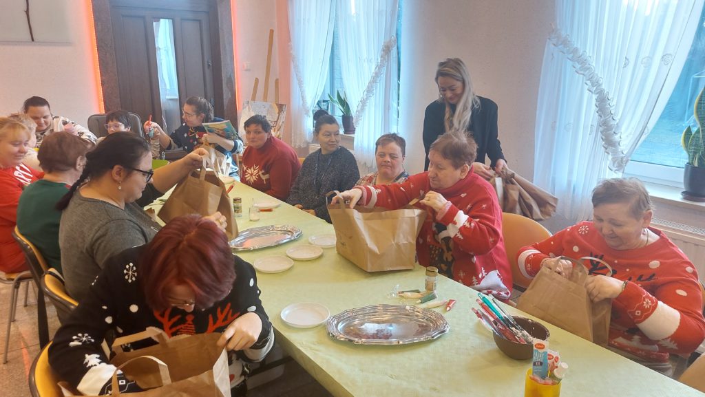 Grupa mieszkanek siedząca przy stołach na sali integracyjno - terapeutycznej ogląda z zainteresowaniem prezentowe papierowe torebki podarowane przez gości z restauracji z mąki. W tle pozostałym mieszkankom nasz gość rozdaje torebki prezentowe 