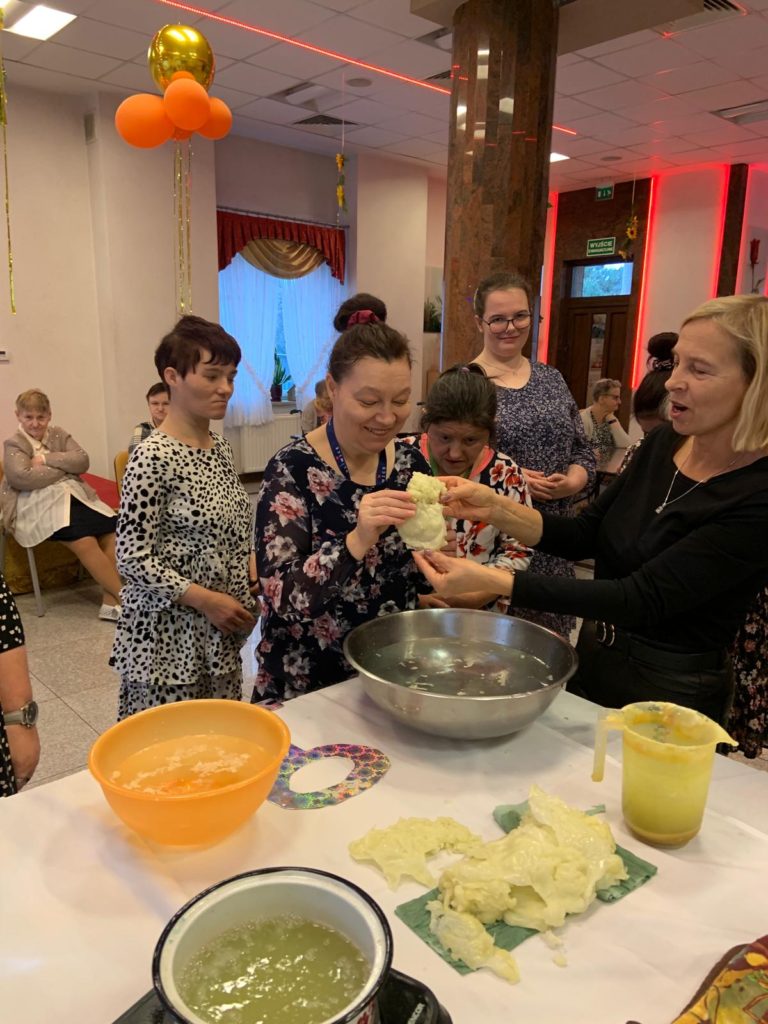 Roześmiana mieszkanka wraz z terapeutką wpatrzone w kształt przelanego wosku nad miską z wodą. W tle kilka mieszkanek wpatrzonych w kształt wosku