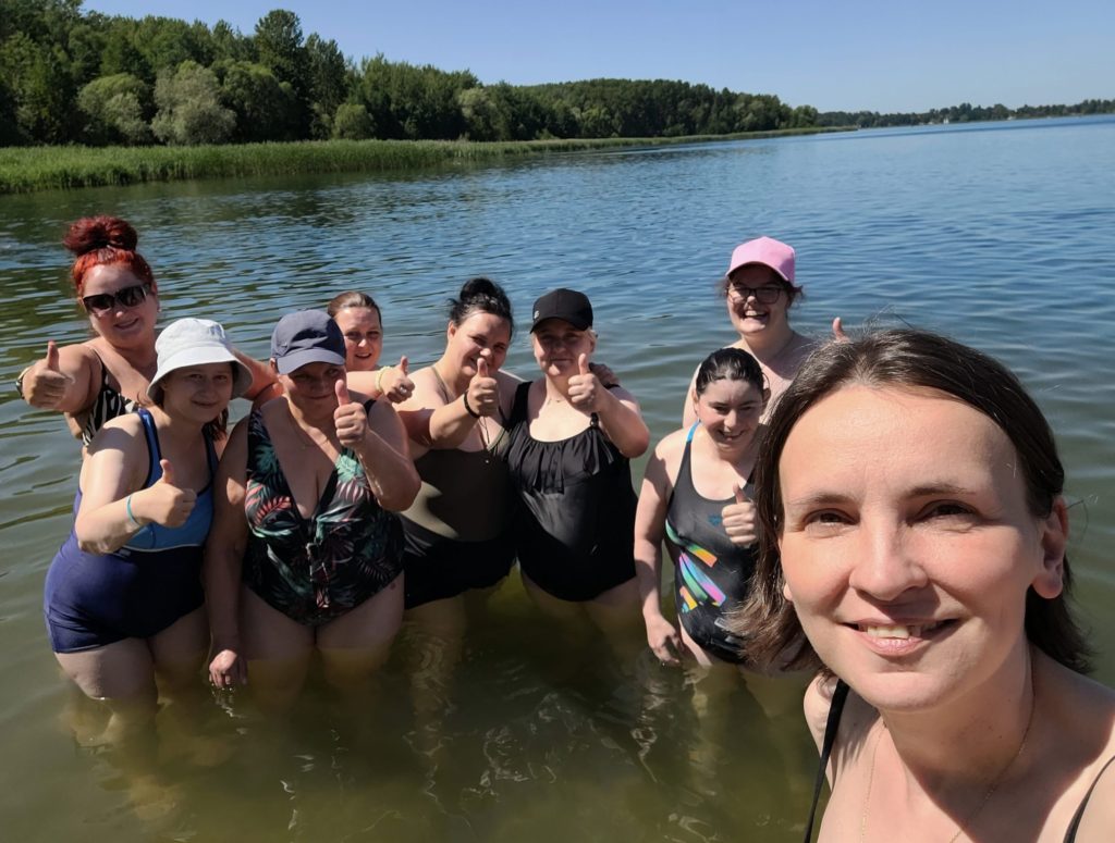 Zdjęcie selfie roześmianej grupy mieszkanek z paniami z personelu w wodzie na tle pięknego krajobrazu. Wszyscy mają kciuki w górę