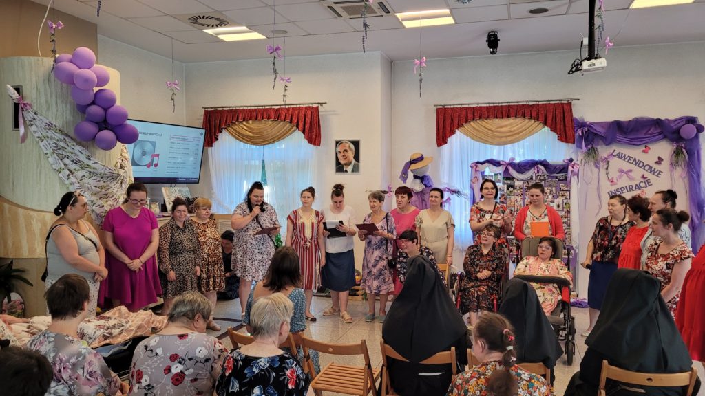 Występ artystyczny mieszkanek dla siostry dyrektor na sali integracyjno - terapeutycznej. Na pierszym planie widać siedzącą oglądającą publiczność, w tle stojące mieszkanki, które występują