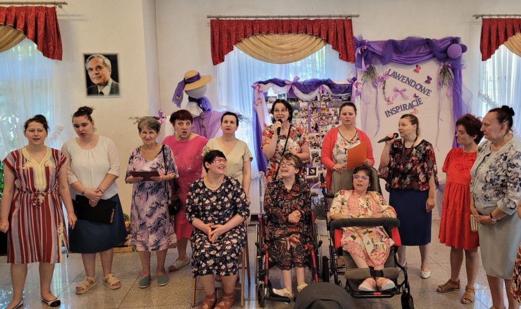 Uśmiechnięta grupa występujących mieszkanek na sali integracyjno - terapeutycznej, dwie z nich trzymają w ręku mikrofon. W grupie występujących są dwie osoby siedzące na wózku inwalidzkim, obok nich osoba siedząca na krześle