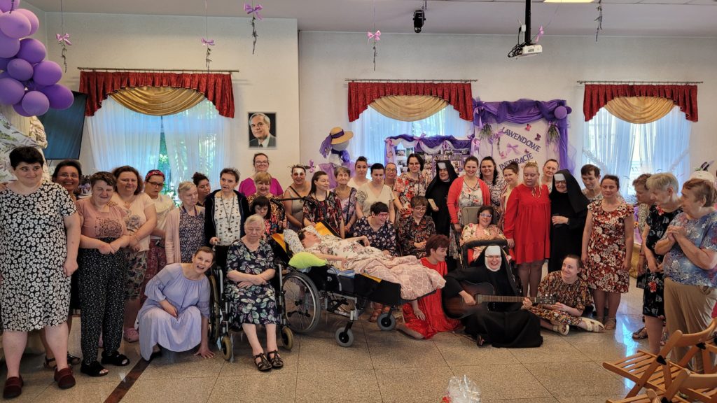 Wspólne zdjęcie roześmianych uczestniczek imieninowego spotkania na sali integracyjno - terapeutycznej. W pierwszym rzędzie kuca kilka mieszkanek oraz siostra albertynka z gitarą, pięć osób siedzi na wózku inwalidzkim