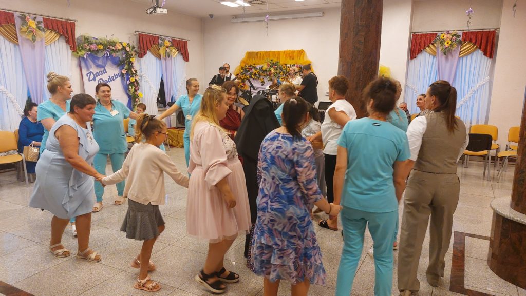 Zabawa taneczna na sali integracyjno - terapeutycznej. Mieszkanki wraz z paniami z personelu tańczą w kółeczku, w tle widoczny zespół muzyczny