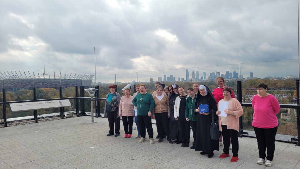 Roześmiana grupa mieszkanek wraz z siostrami Albertynkami stojąc pozuje do zdjecia na tarasie widokowym na dachu fabryki czekolady. W tle stadion narodowy i budynki Warszawy