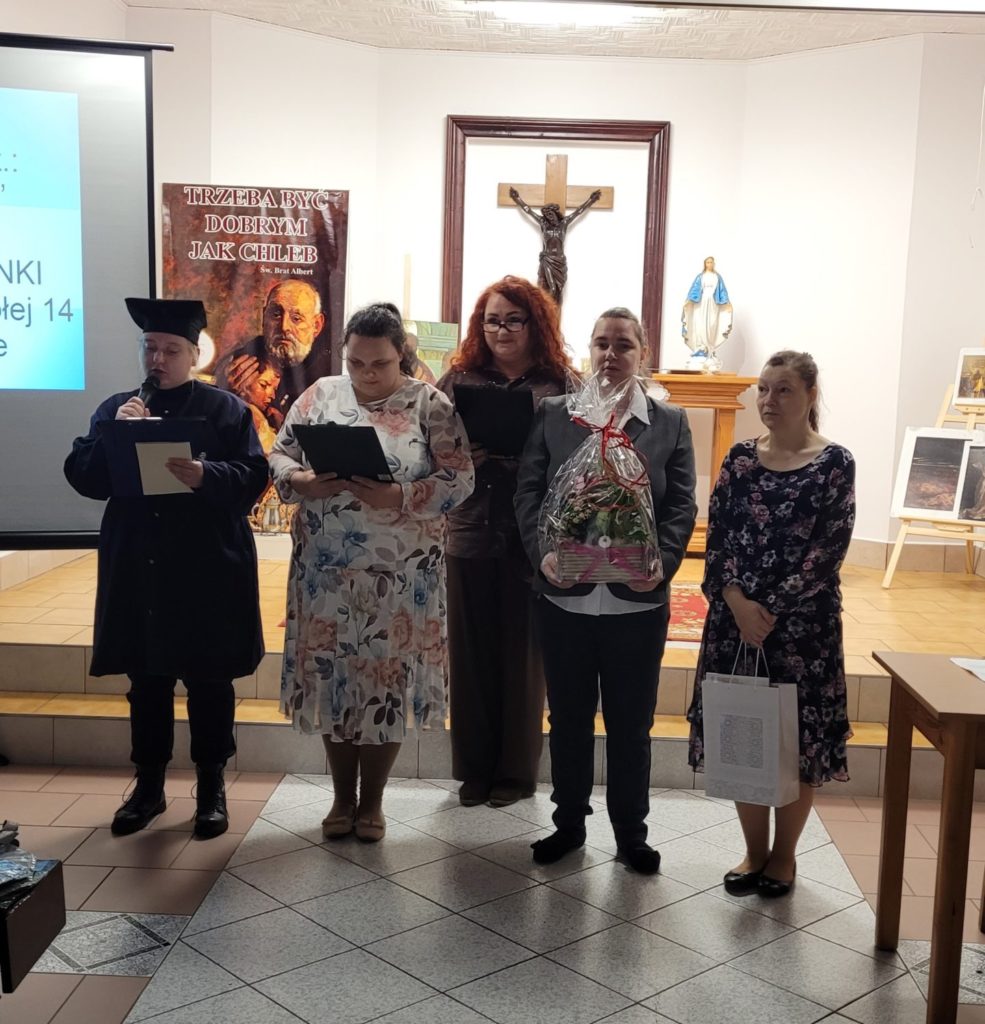 Grupa czterech mieszkanek stojąc składa życzenia siostrze solenizantce, dwie z nich mają w rękach kwiaty i prezent, pozostałe osoby trzymają podładkę z tekstem.. W środku między nimi stoi opiekunka grupy, w tle krzyż i plakat ze świętym bratem Albertem.