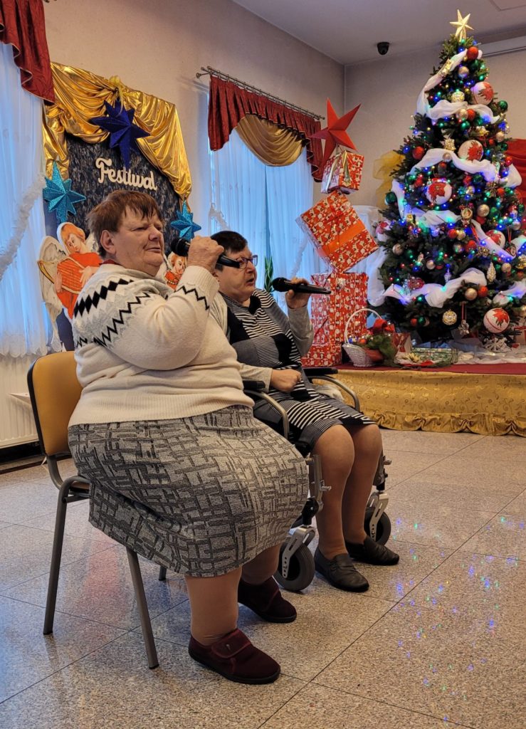 Dwie mieszkanki siedzą na krzesełkach na sali integracyjno - terapeutycznej podczas swojego występu na festiwalu kolęd. Obie trzymają w ręku mikrofony, w tle jest choinka