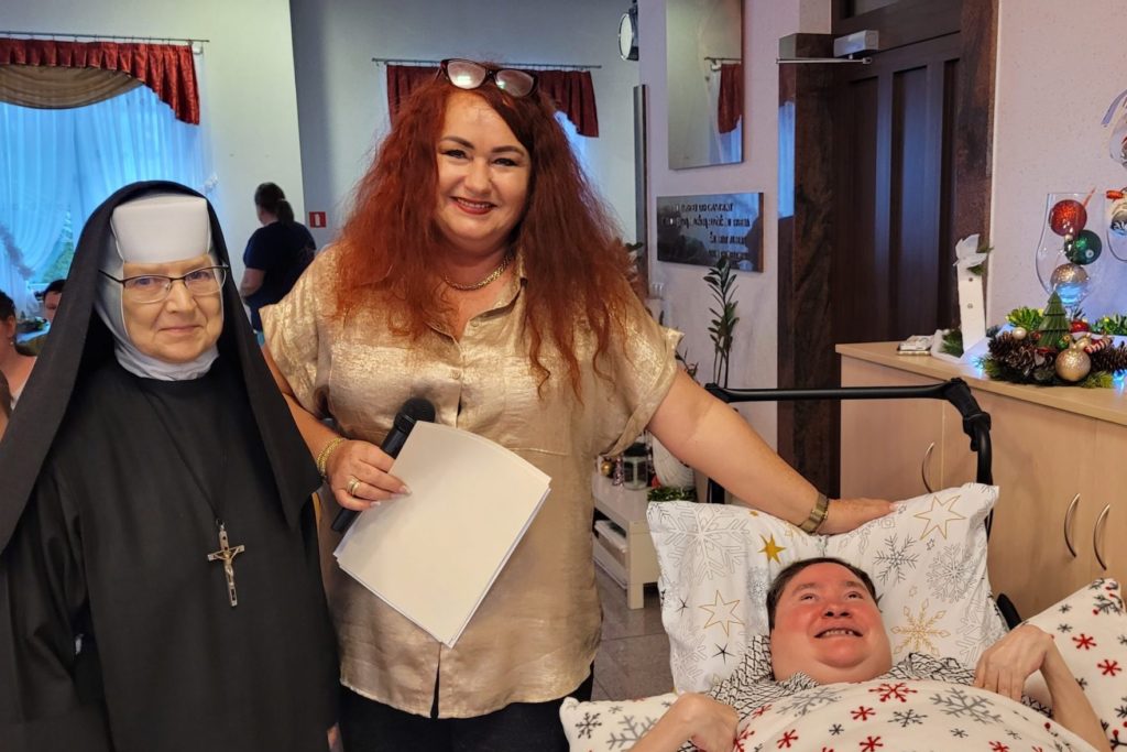 Uśmiechnięte dwie osoby z jury, w tym siostra Albertynka, stojąc pozują z leżącą uczestniczką festiwalu kolęd. 