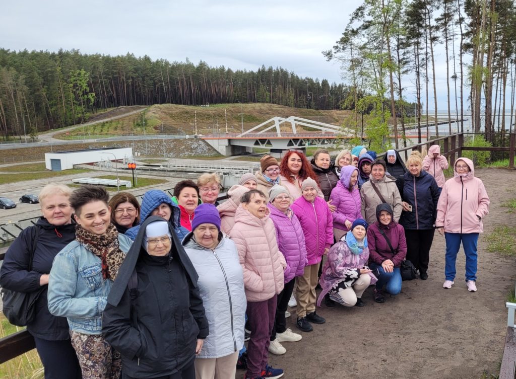 Wspólne zdjęcie uśmiechniętych stojących uczestników wycieczki na tle Mierzei Wiślanej. Dwie osoby kucają przed grupą pozując do zdjęcia