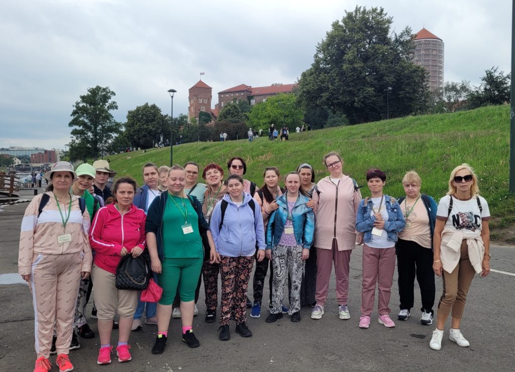 Grupa mieszkanek wraz z jedną z opiekunek grupy pozuje do zdjęcia stojąc na deptaku przy Wiśle - w tle widać Wawel