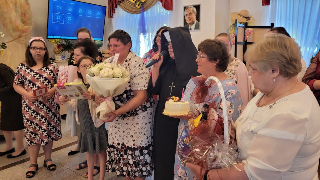 Na sali integracyjno - terapeutycznej stoją mieszkanki, pośrodku stoi siostra dyrektor z mikrofonem w ręku. Mieszkanki kolejno mają w ręku upominki - czekoladki, kwiaty, tort, kosz prezentowy